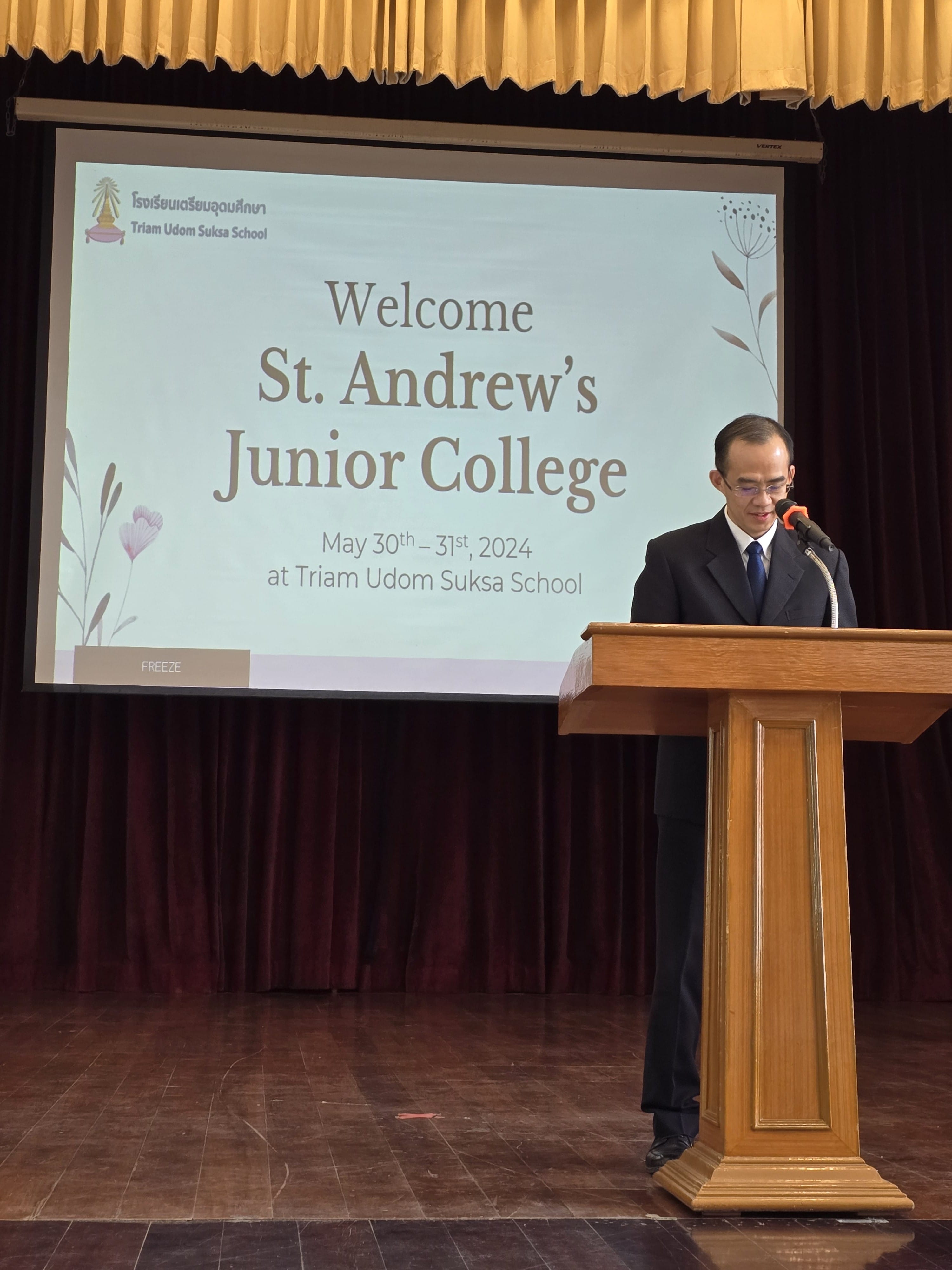A speaker stands at a podium beside a projected welcome slide reading 'Welcome St Andrew's Junior College, May 30th–31st, 2024 at Triam Udom Suksa School'.
